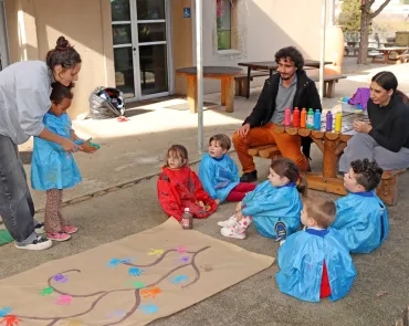 des enfants, accompagnés d'adultes posent leur main pleine de peinture sur une fresque en forme d'arbre pour former des feuilles de mains