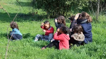 des enfants et leur animatrice sont asssis dans l'herbe et regardent au travers de jumelles