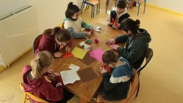 Cinq enfants et leur animateur plient du papier sur une table
