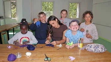 Un groupe d'enfants et leur animatrice sont en salle en train de tisser de la laine