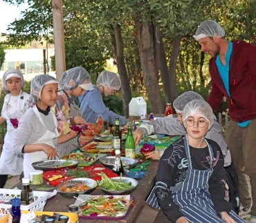 Des enfants avec tabliers et charlottes cuisinent des légumes sur des tables extérieures 