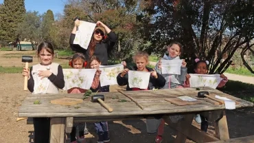 Sur une table en bois de pique-nique, des enfants et leur animatrice montrent des maillets et des morceaux de tissus imprimés