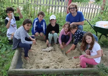 un groupe d'enfants et leur animateur ont les pieds et les mains dans un bac à sable