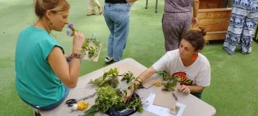 à gauche, une femme assie sur un bout de table sent une fleur. En face, une autre femme réalise un tissage nature