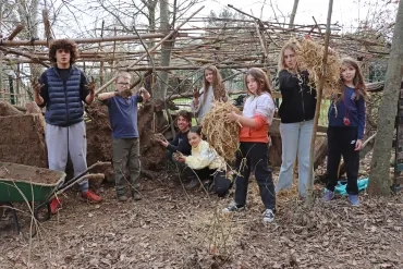 un groupe d'enfants et leurs animateurs montrent de la paille et de la boue devant une cabane
