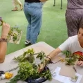 à gauche, une femme assie sur un bout de table sent une fleur. En face, une autre femme réalise un tissage nature